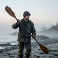 Portrait of a young man in a woolly hat holding a kayak paddle on a misty shore