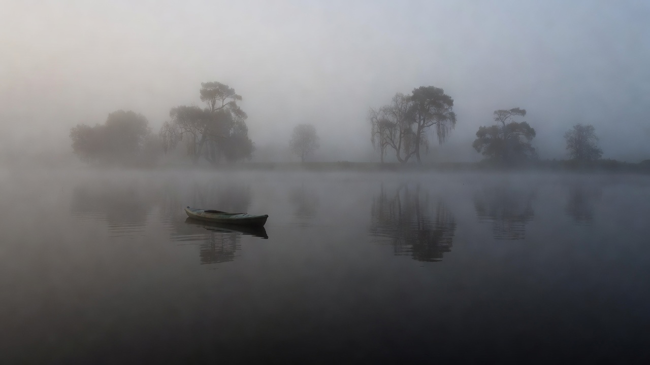 A kayak gliding across black, glassy water through dense dawn fog with faint silhouettes of trees